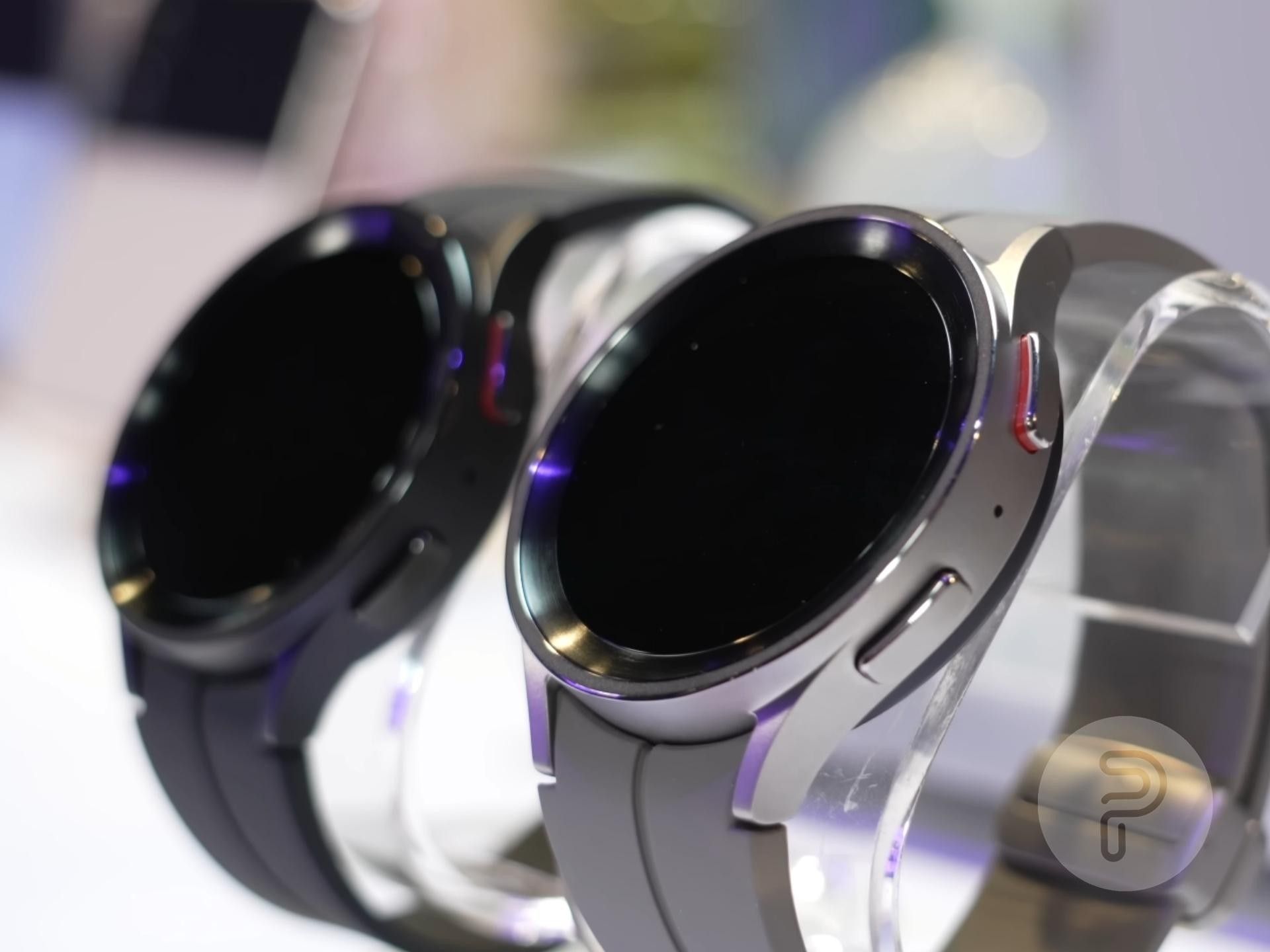 Arka planda Siyah Titanyum varyantı ile Gri Titanyum Galaxy Watch 5 Pro;  her ikisi de bir saat tutucuya yerleştirilmiştir